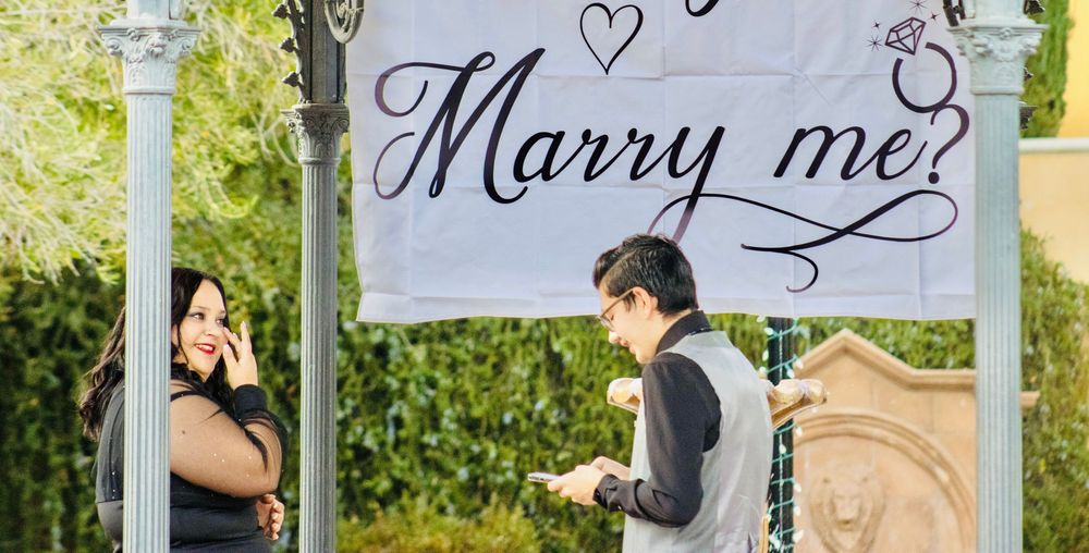 Couple standing under a Will You Marry Me banner during a marriage proposal in an outdoor gazebo.