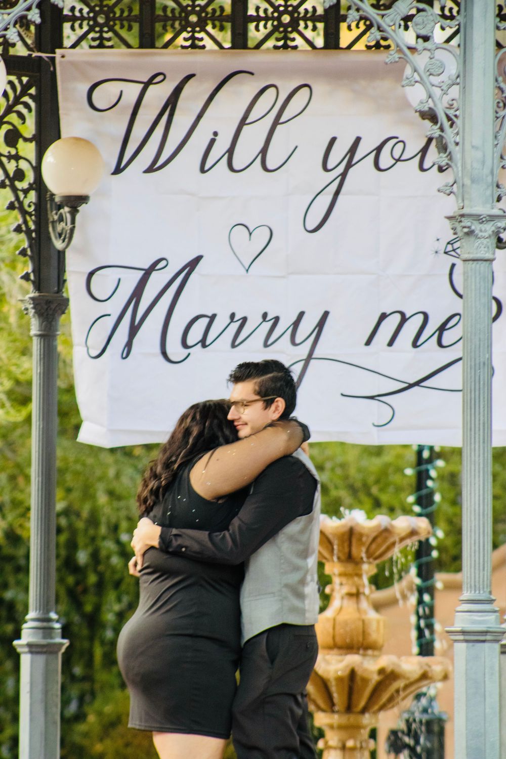 Couple embracing under a Will You Marry Me banner in an ornate gazebo with fountain.