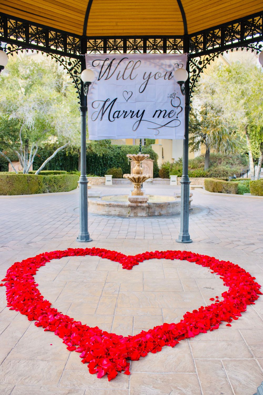 Romantic marriage proposal setup with red rose petal heart and Will You Marry Me banner in gazebo.