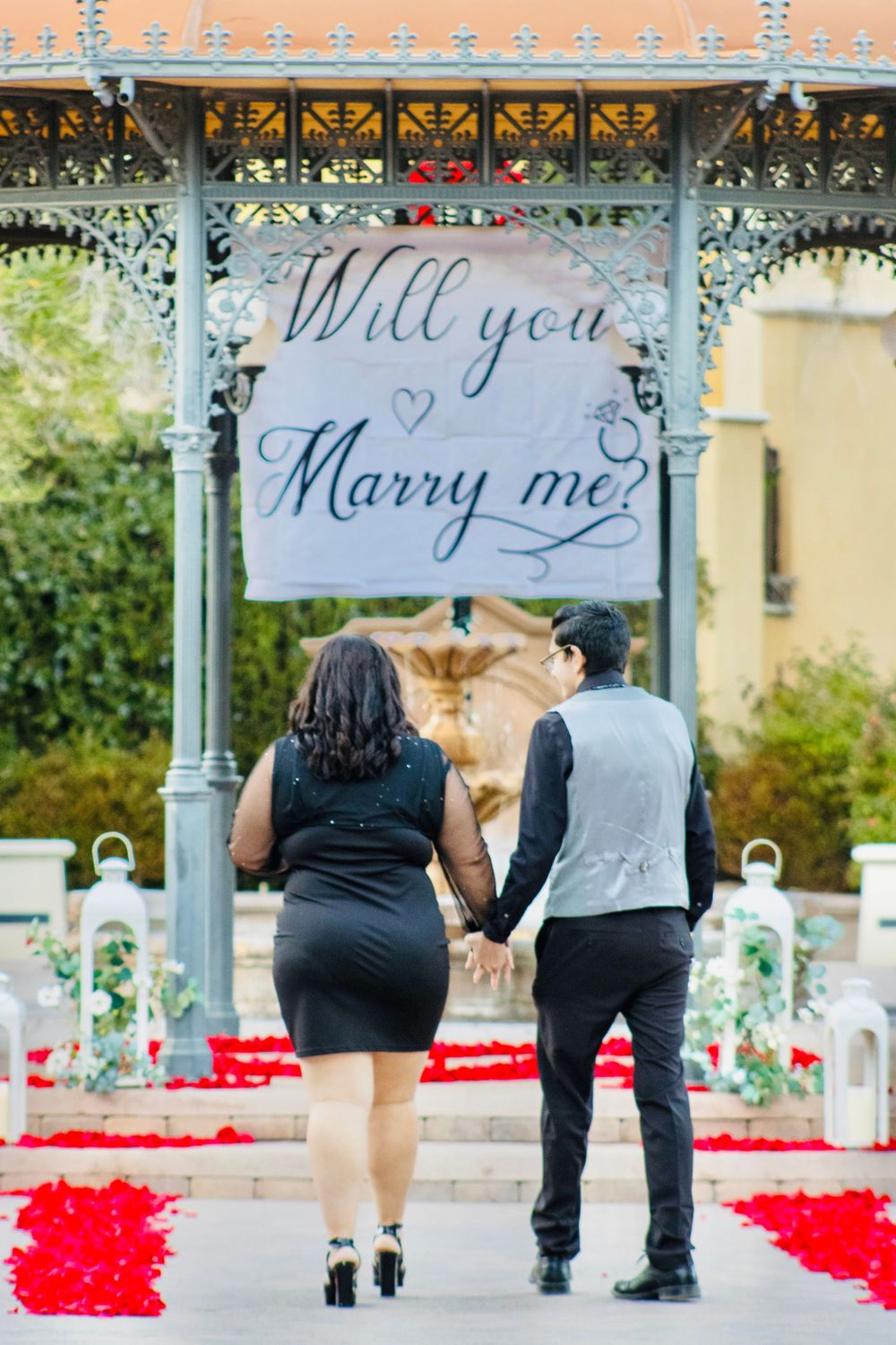Couple holding hands walking toward a Will You Marry Me banner in a rose petal decorated gazebo.