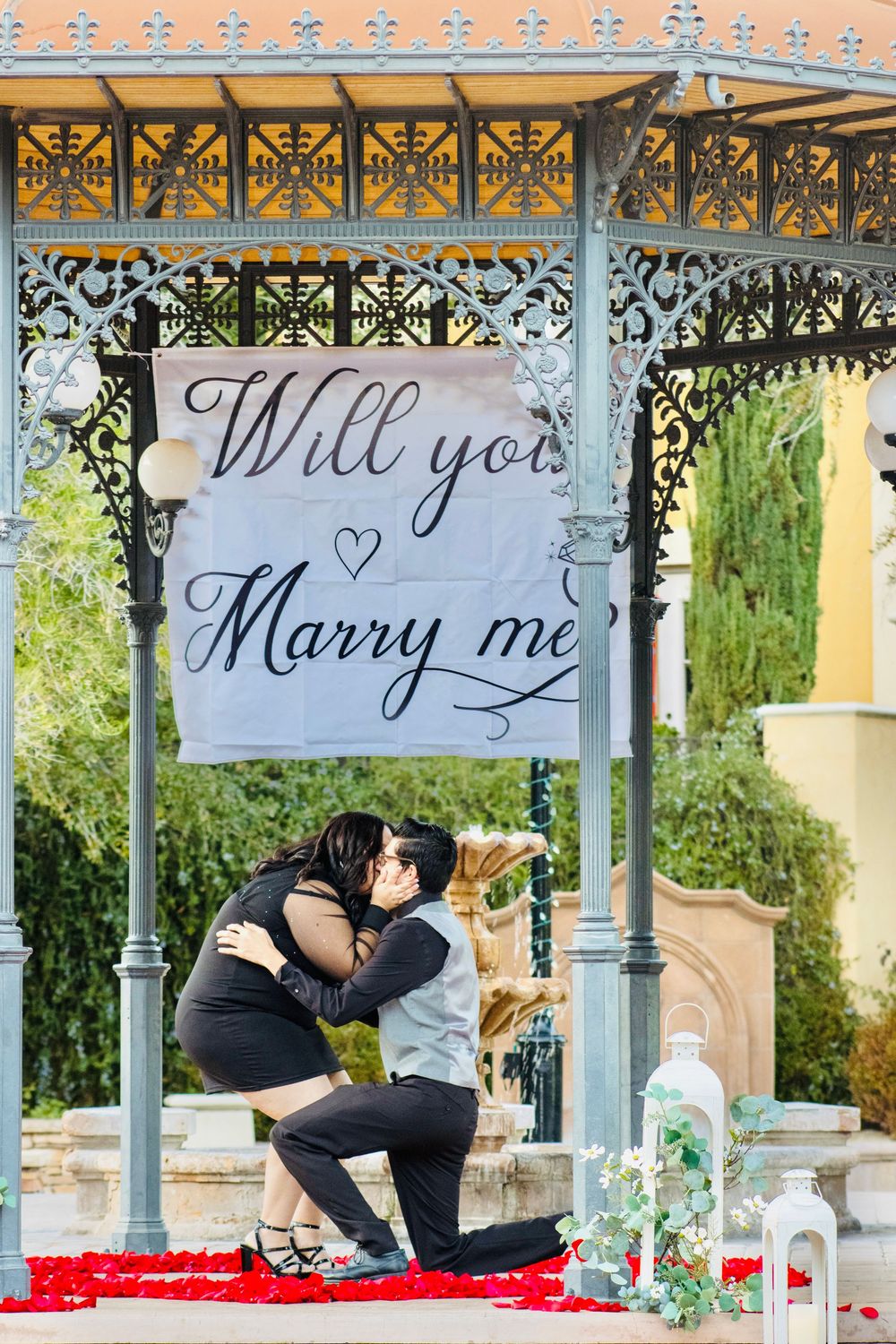 Man proposes on one knee to woman under Will You Marry Me banner in decorated gazebo.