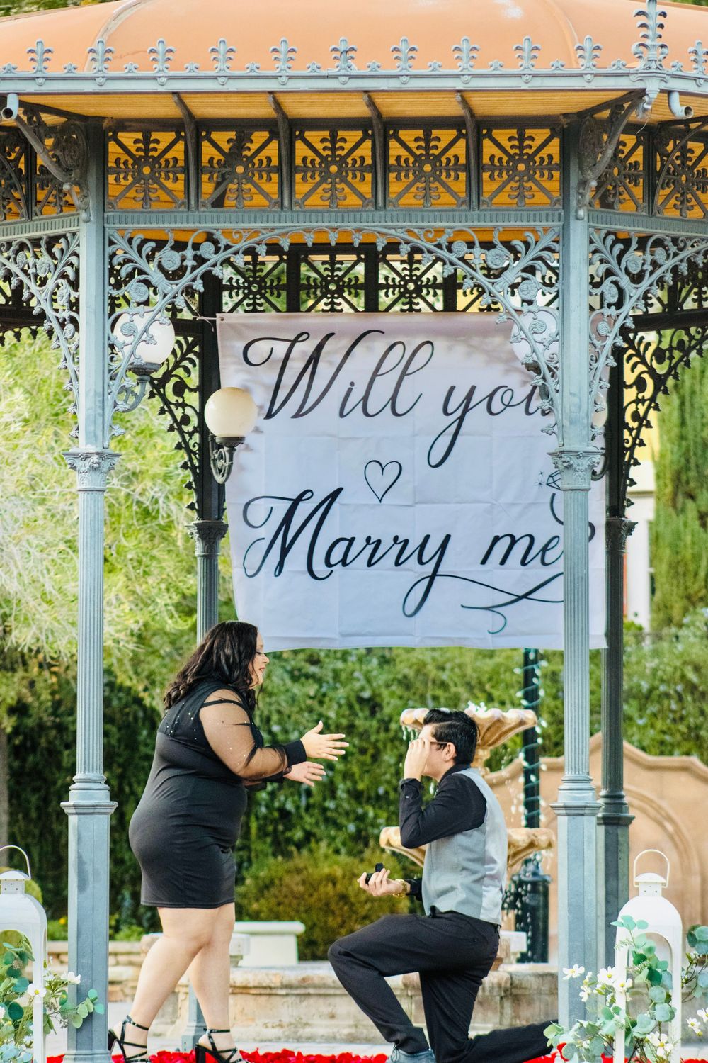 Romantic marriage proposal under an ornate gazebo with Will You Marry Me banner and rose petals.