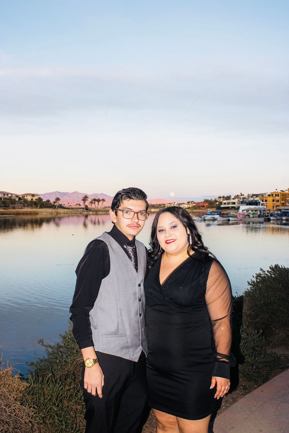 Engaged couple standing together by a scenic lake at dusk with mountains in the background.