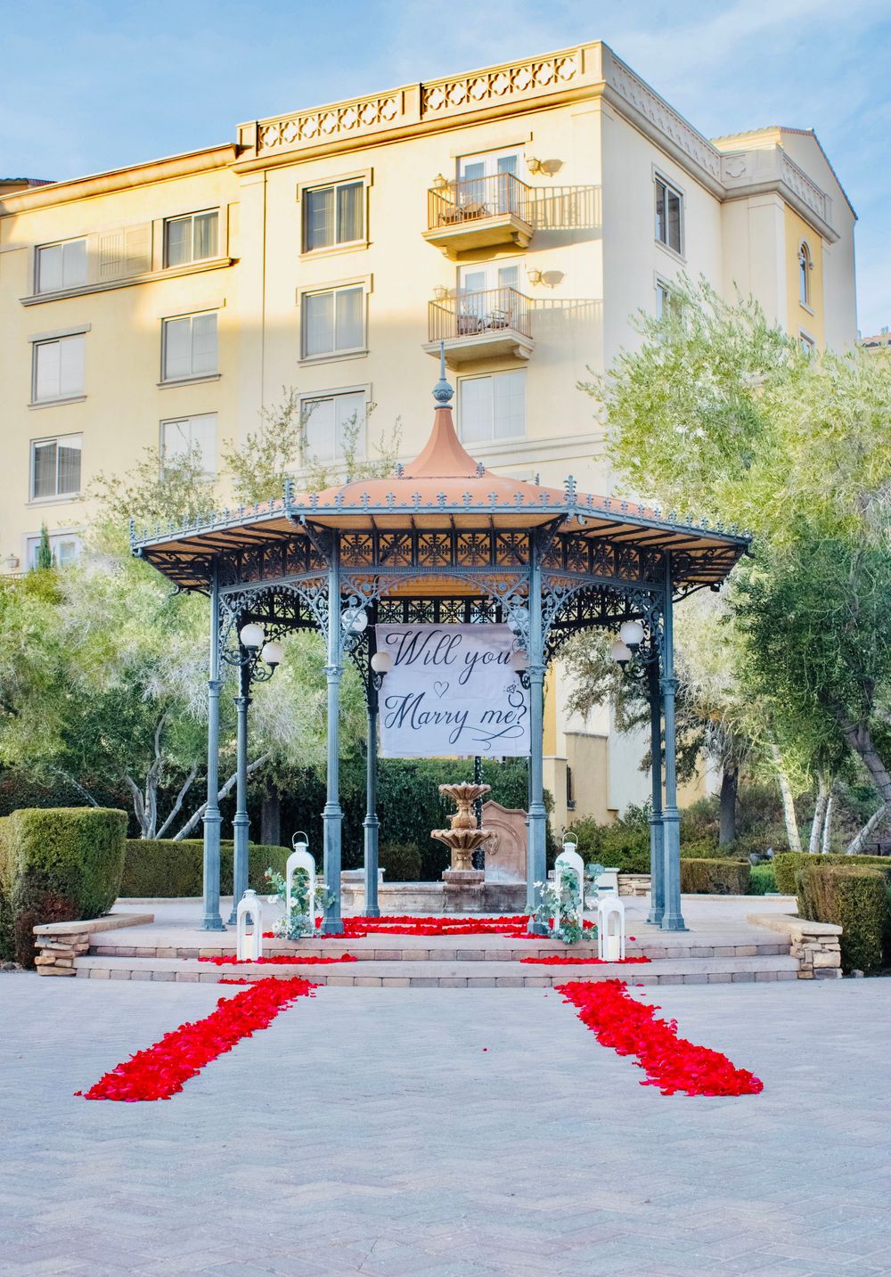 Ornate outdoor gazebo with red rose petal path and Will You Marry Me banner at a hotel.
