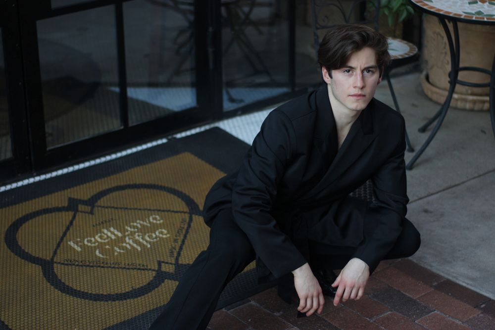 Young man in black suit sitting on brick floor near Fearless Coffee entrance mat.
