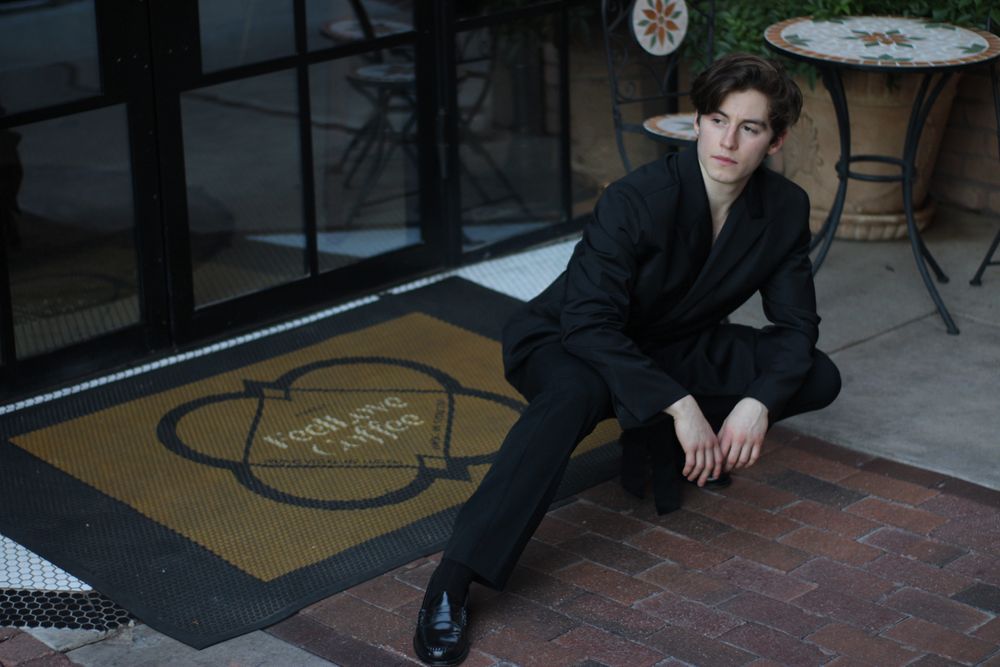 Stylish young man in black blazer posing on brick floor outside coffee shop.