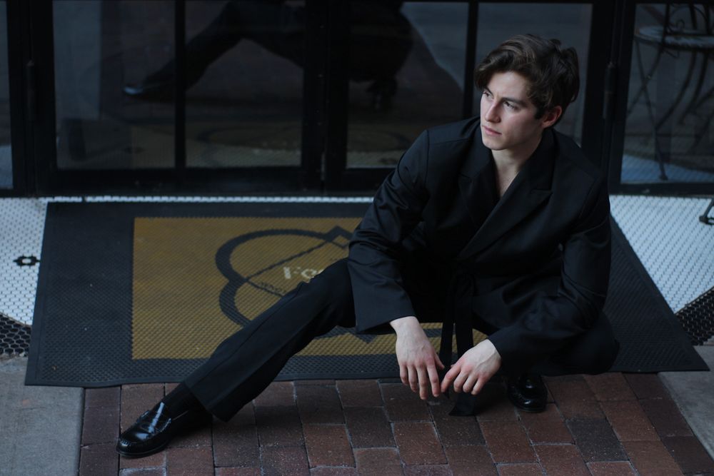 Young man in black suit sitting casually on brick pavement near glass door.