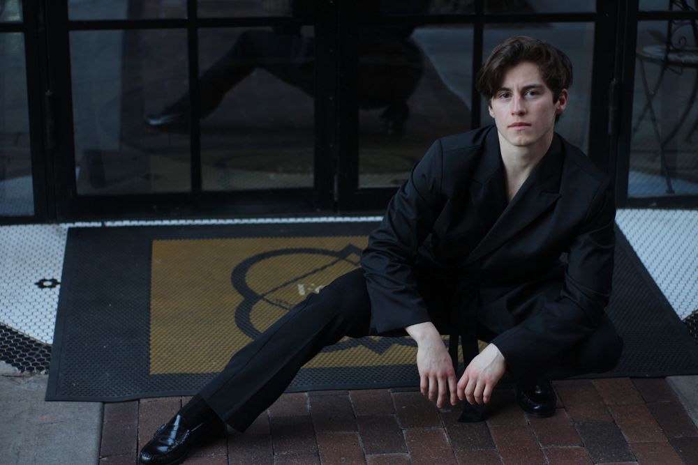 Fashionable young man in black outfit seated on brick floor near entrance.