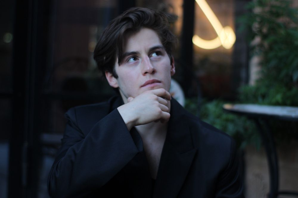 Thoughtful young man in black blazer with hand on chin at outdoor cafe.