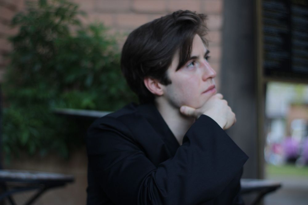 Young man in black jacket posing pensively with hand under chin outdoors.
