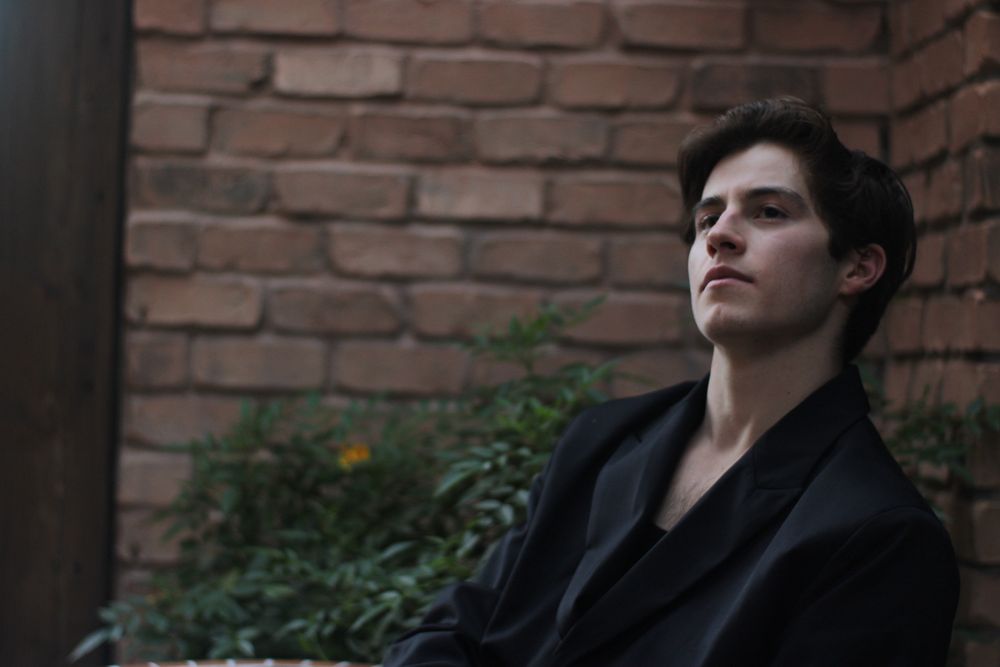 Young man in black jacket sitting pensively against rustic brick wall in moody outdoor setting.