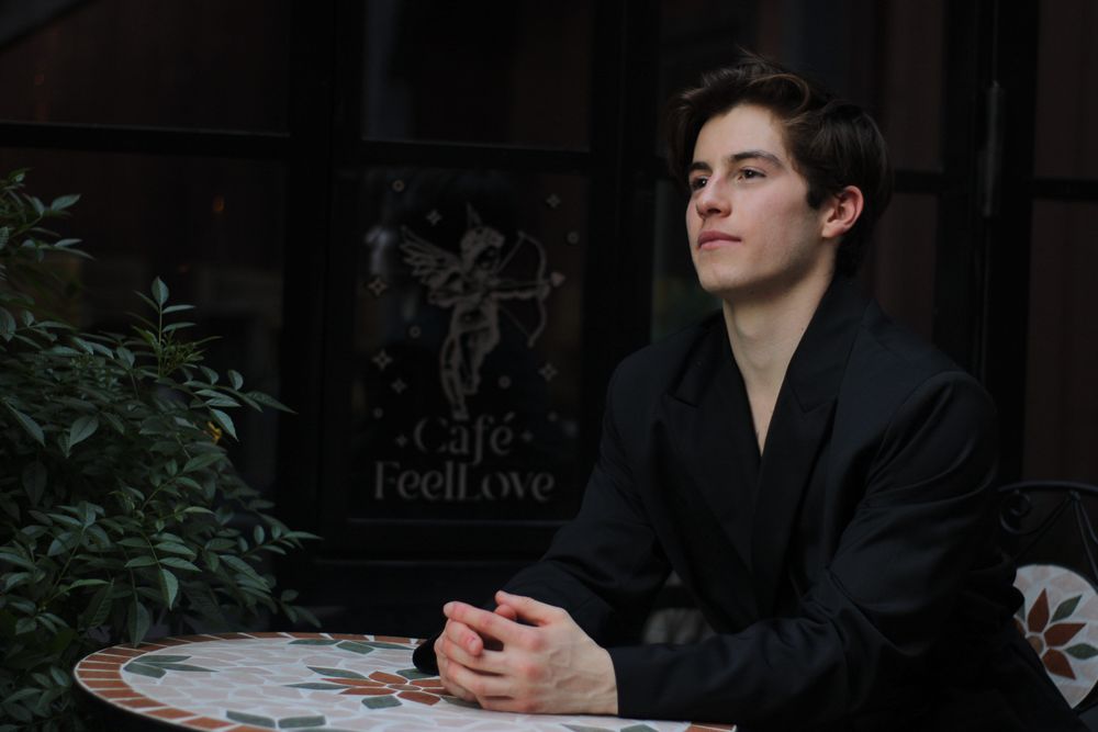 Young man in black jacket sitting at mosaic cafe table near Cafe Feel Love sign outdoors.