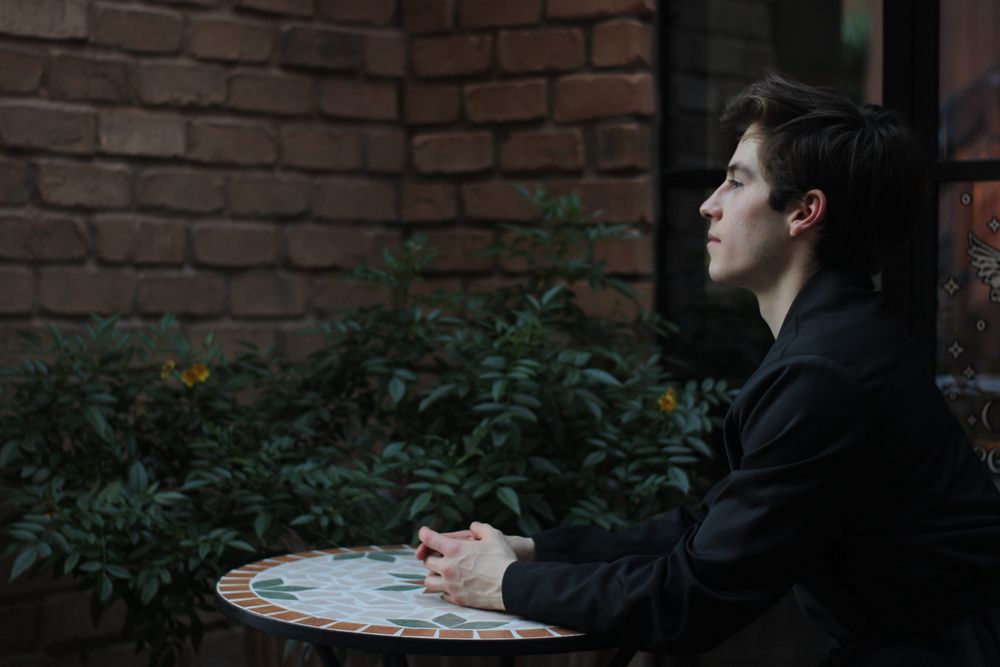 Side profile of young man in black jacket sitting at mosaic table against lush brick wall.