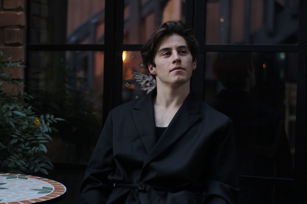 Young man in oversized black belted jacket seated at outdoor cafe table in moody fashion shot.