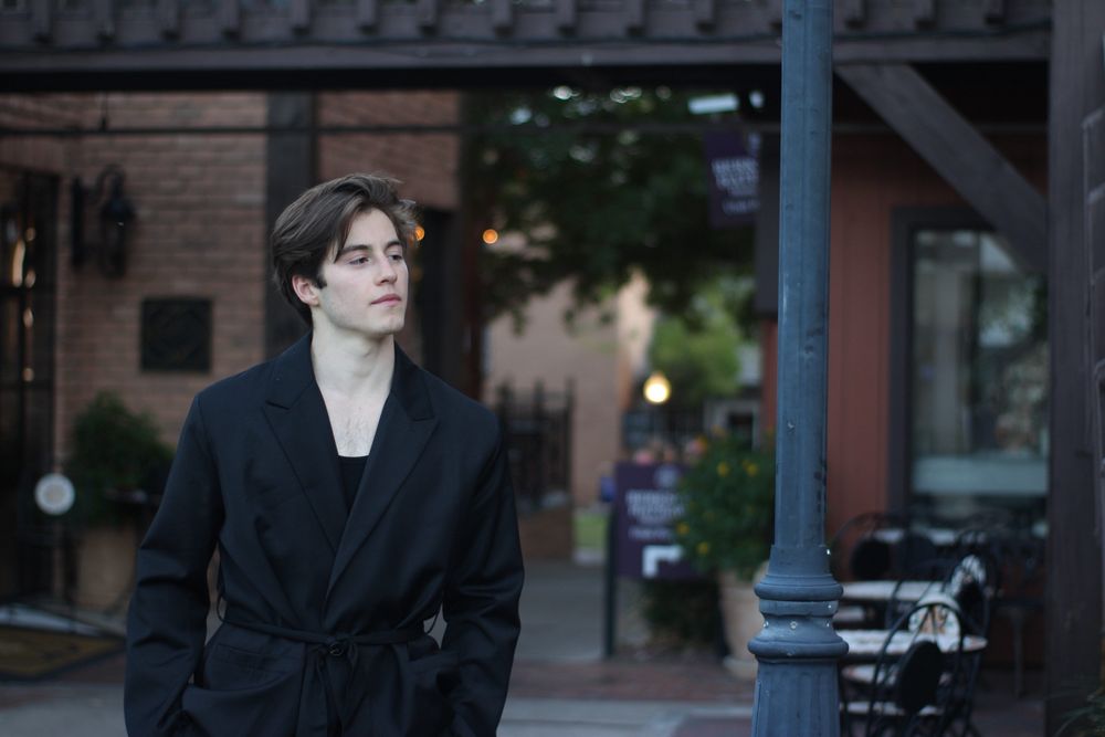Young man in black oversized coat standing near street lamp in urban outdoor fashion editorial.