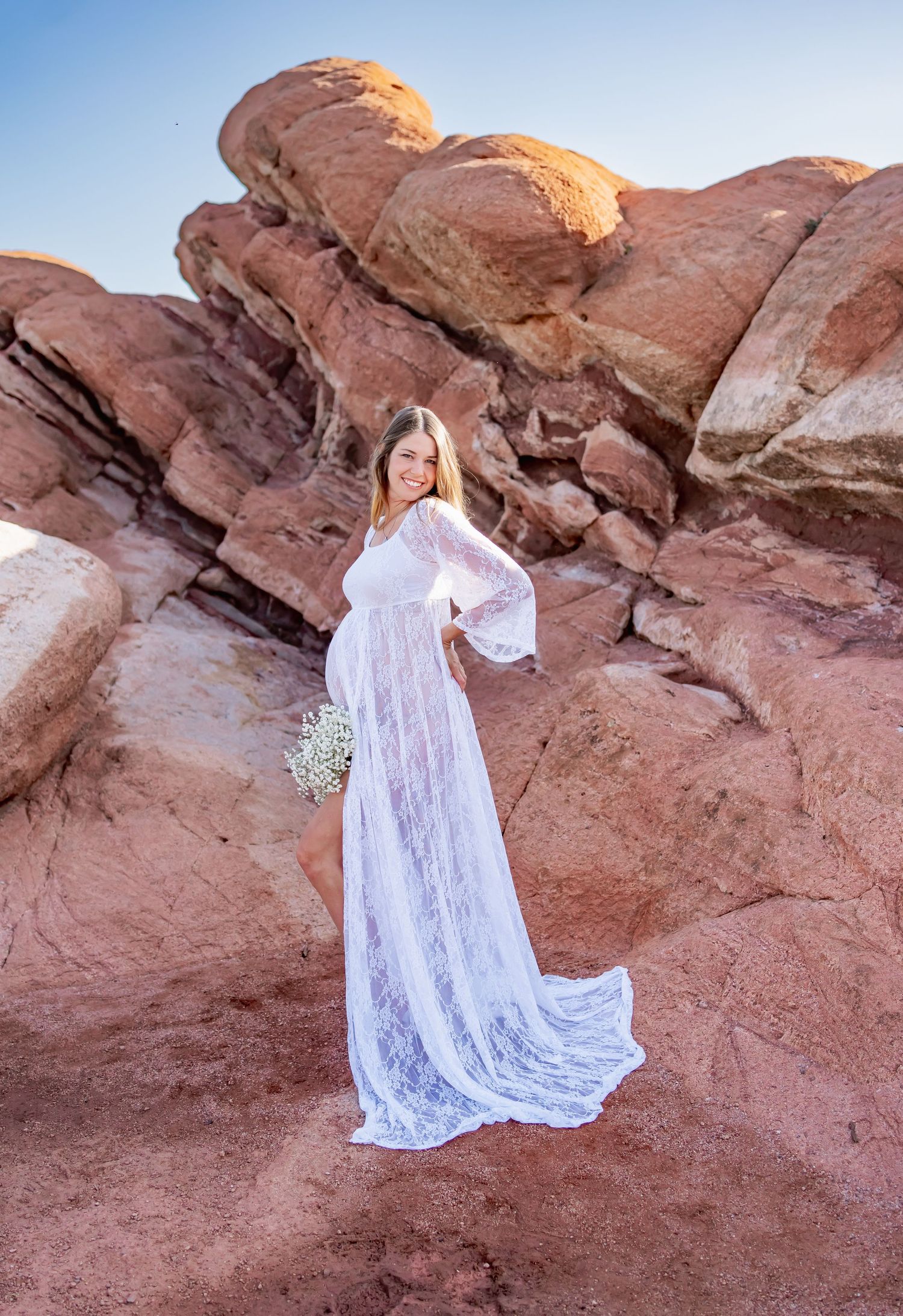 A dramatic maternity photo with white lace dress against red rock formations at sunset.
