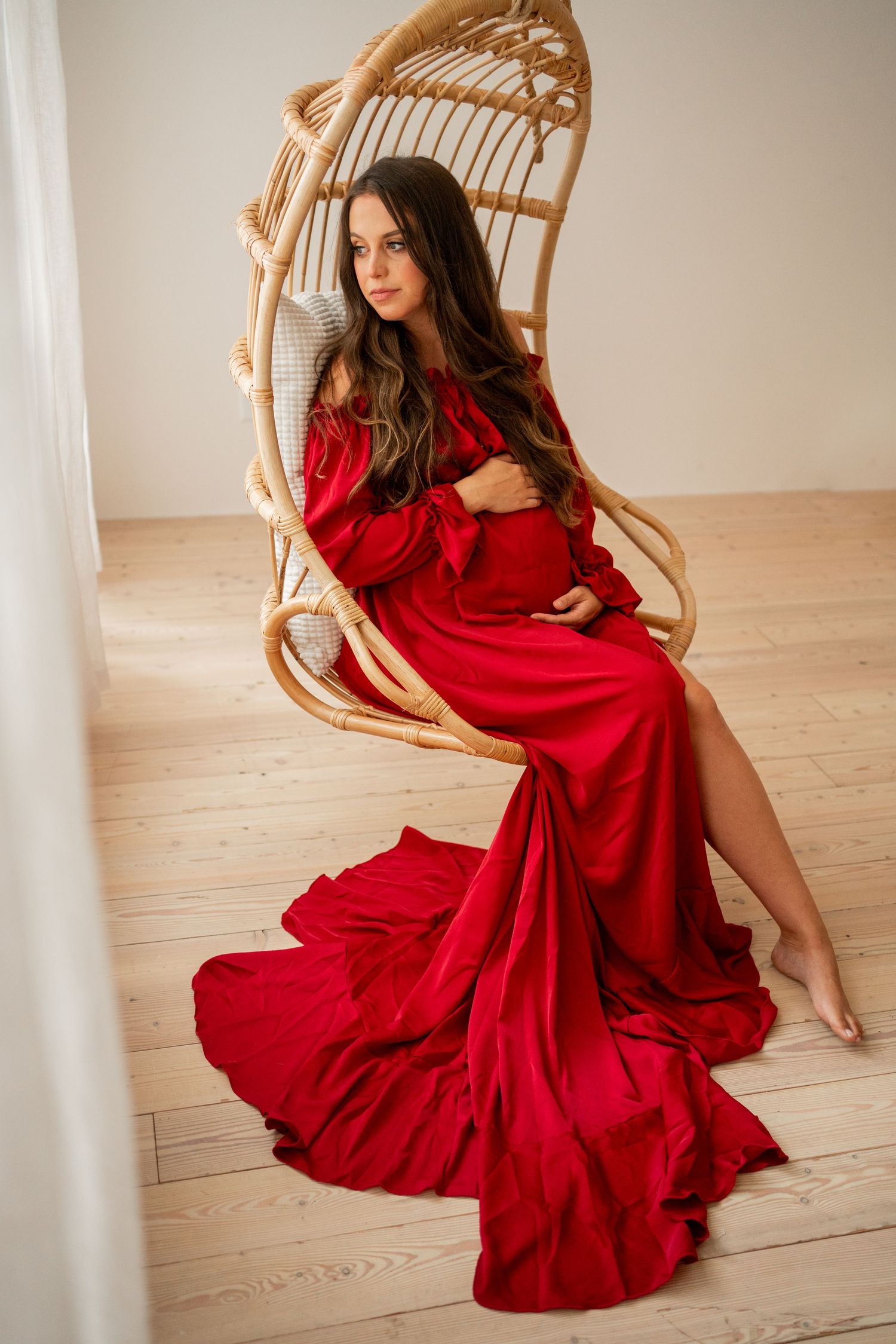 Red maternity gown flowing dramatically around figure seated in rattan peacock chair.