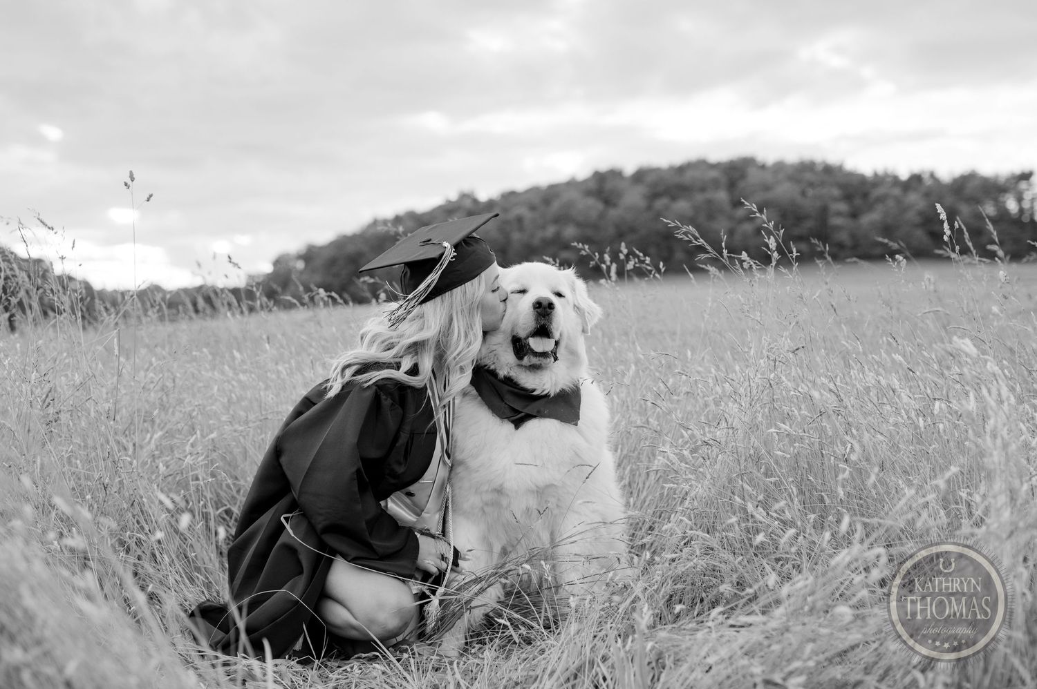 Black and white portrait of a happy dog and graduate sitting in a grassy meadow.