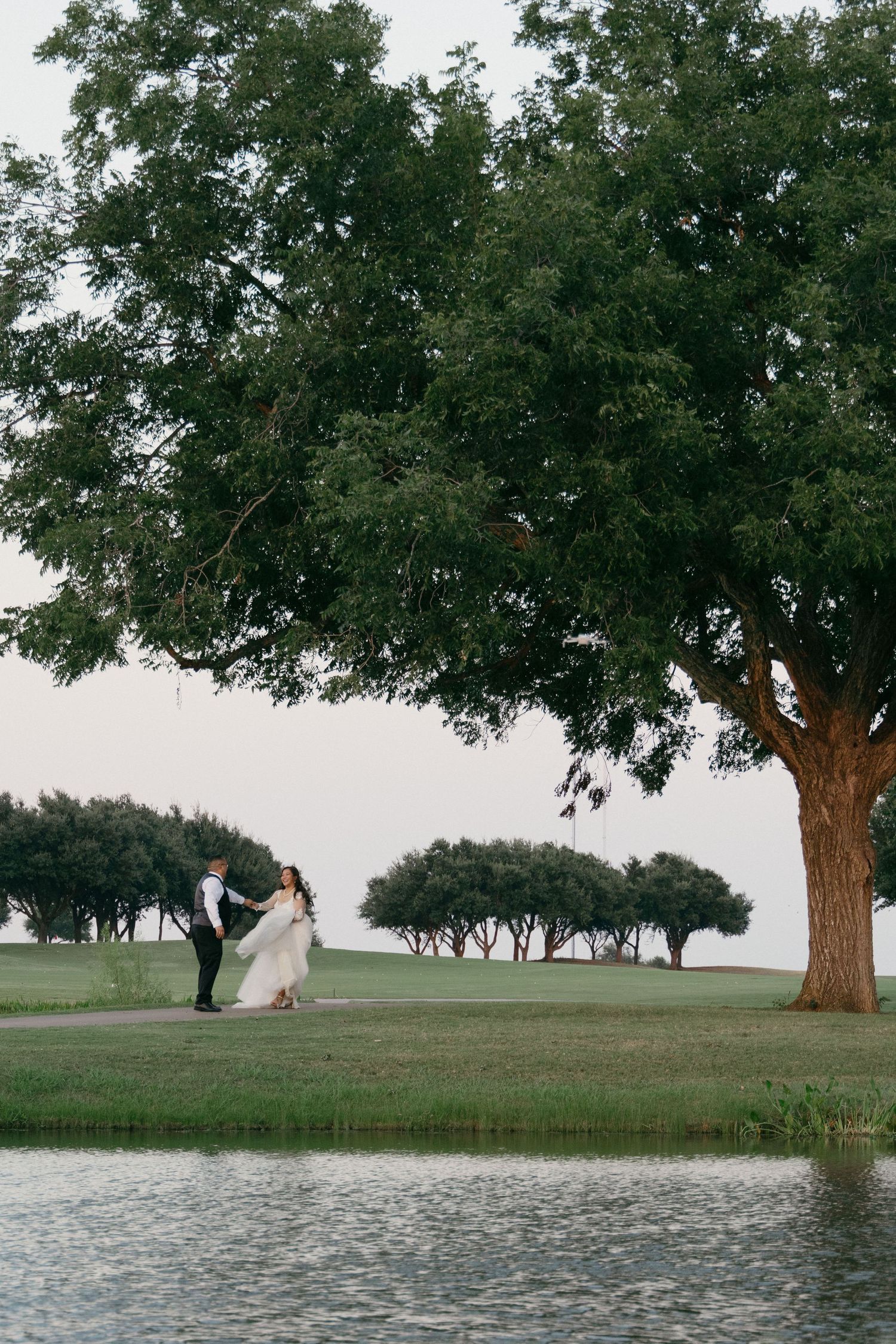 Wedding couple stands near a large tree by a lake in a peaceful park setting.