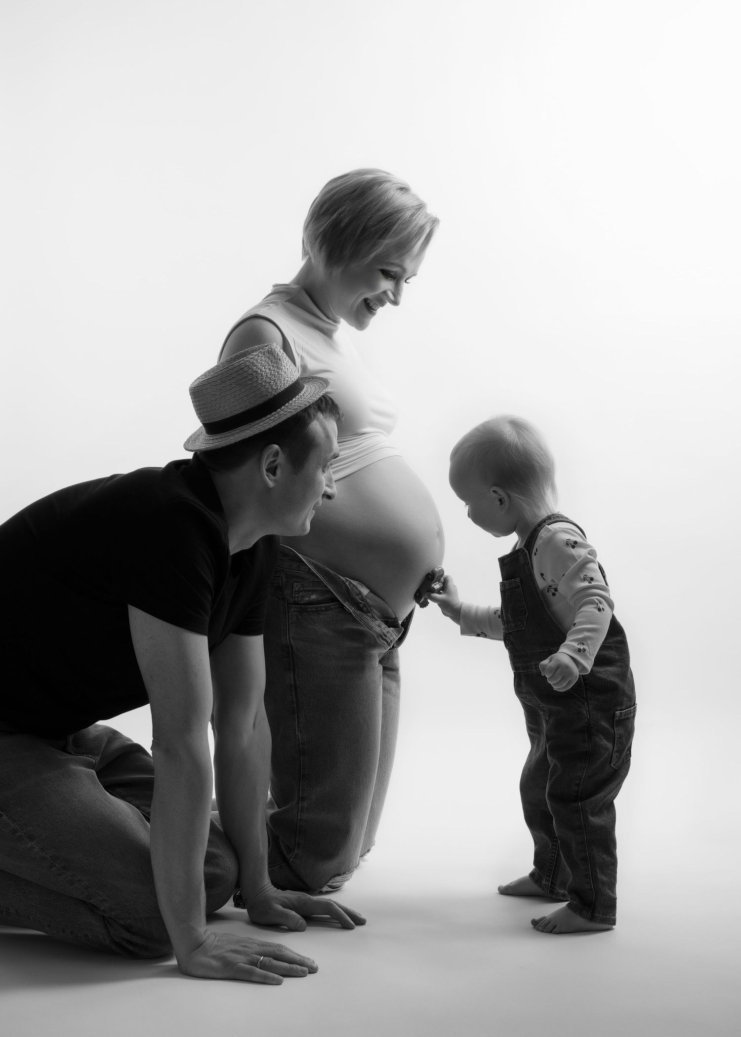 from a maternity photo shoot in studio, a mother kneels down next to her partner + by her child so her belly faces them