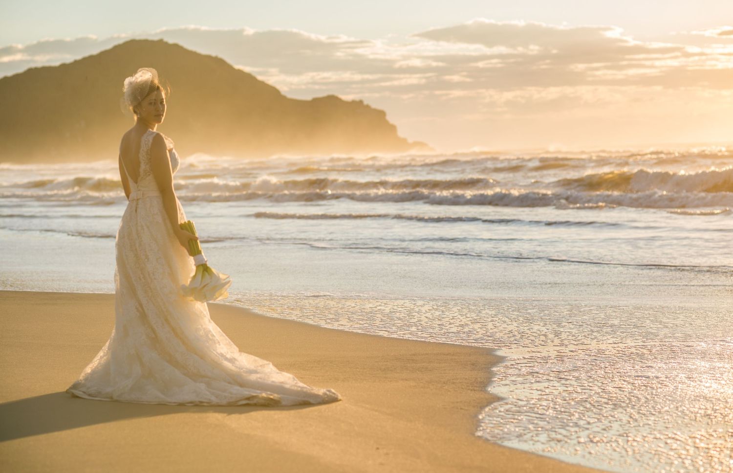 Elegant white wedding dress silhouetted against golden sunset on beach with mountain backdrop.