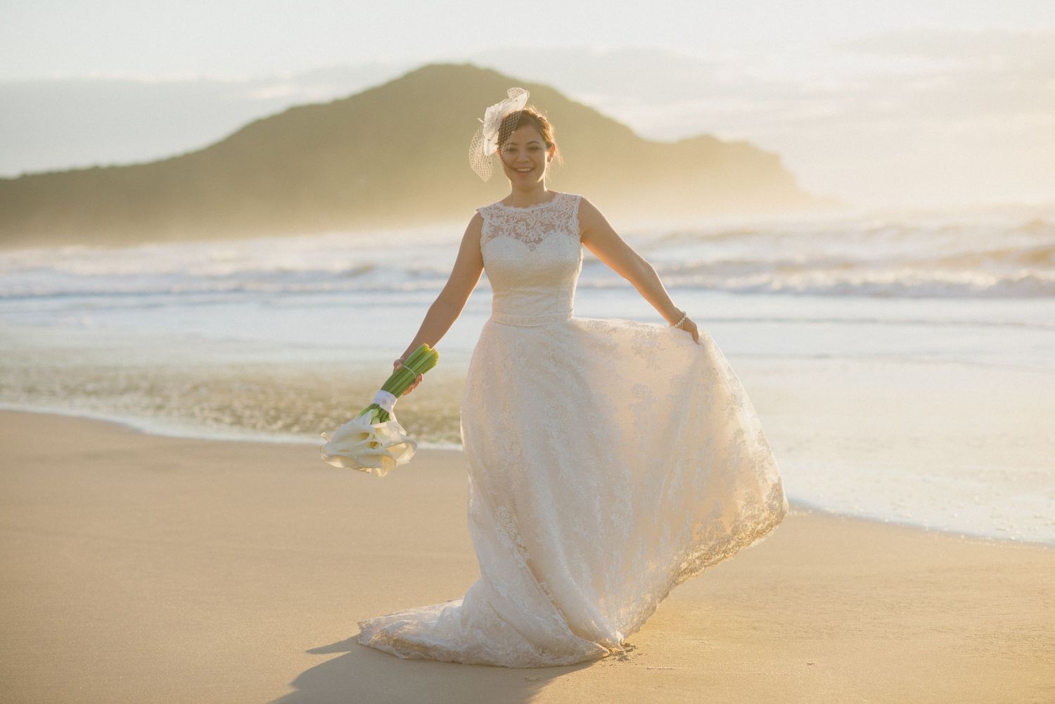 Radiant beach wedding scene with flowing white gown against mountain landscape at sunset.