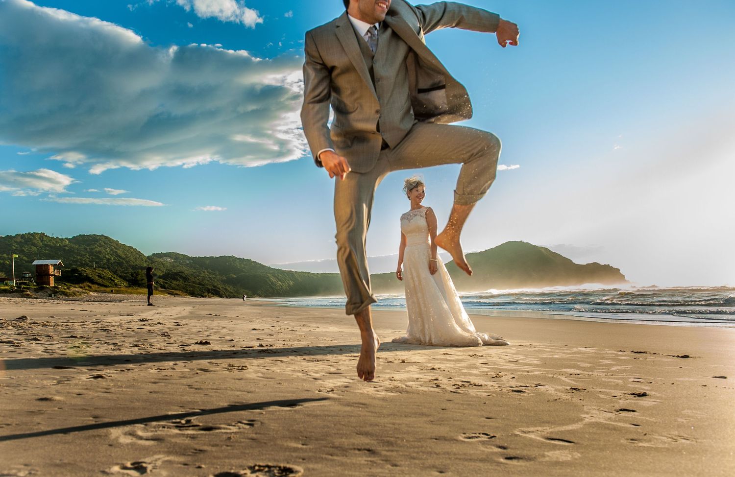 Playful beach wedding photo with groom jumping in suit against coastal landscape.