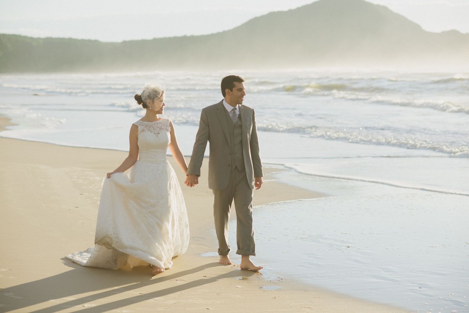 Newlyweds walking hand in hand along beach shoreline at sunset.