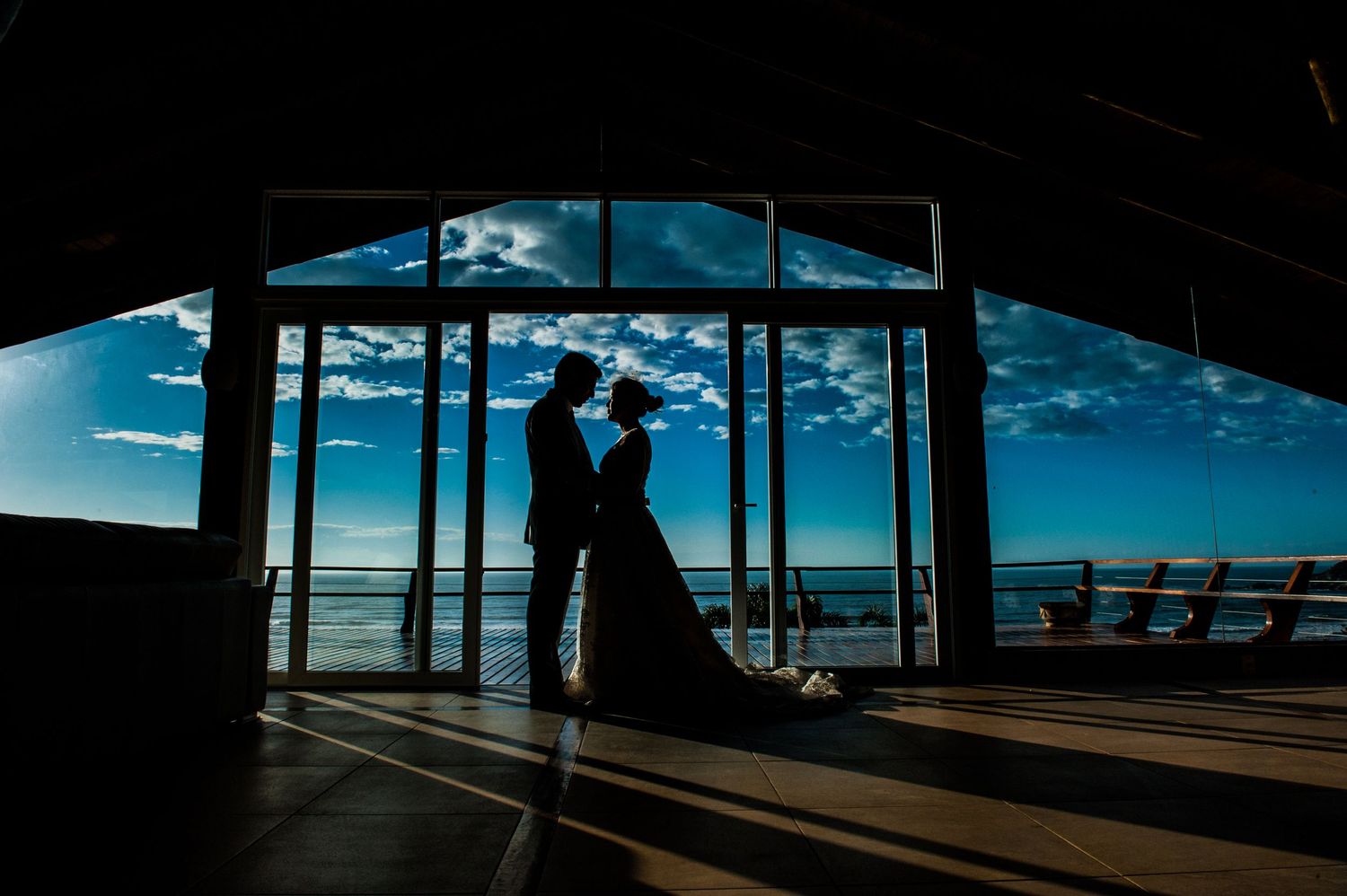 Silhouetted figures in romantic pose against dramatic ocean view through architectural framing at sunset.