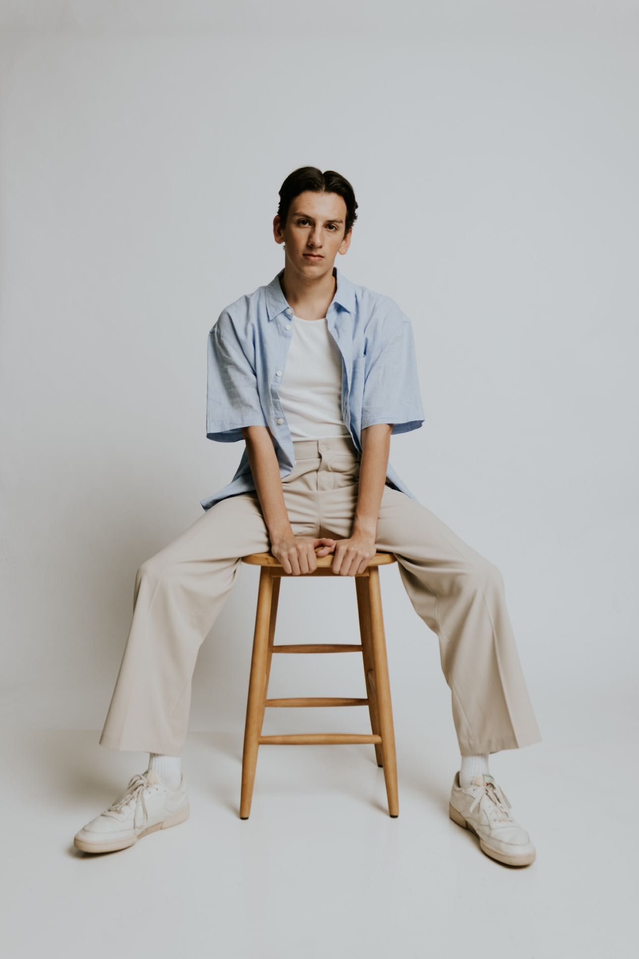 Casual studio portraits on wooden stool wearing light blue shirt and khaki pants.