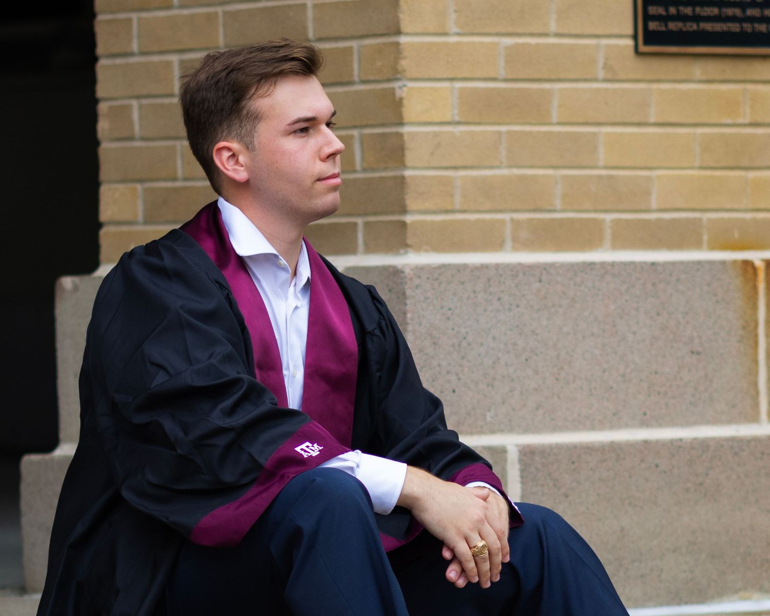 Graduate in traditional academic regalia sitting thoughtfully against brick wall.