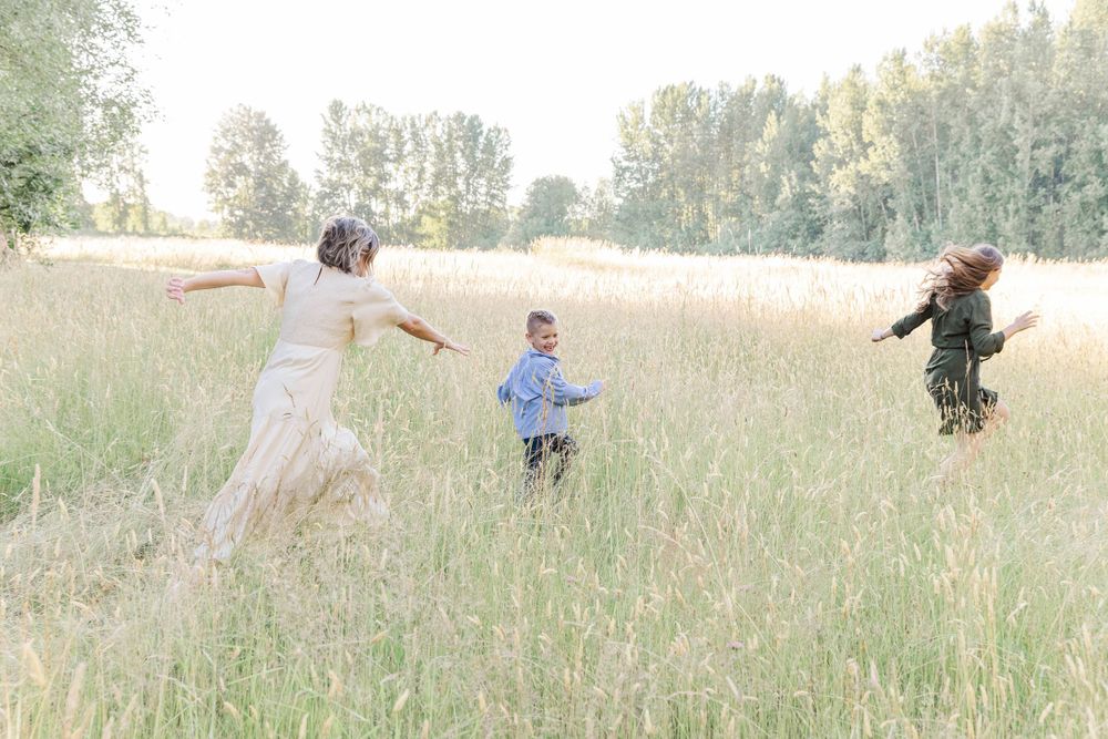 Woman in white dress plays with two children running through a tall grass meadow on a sunny day.