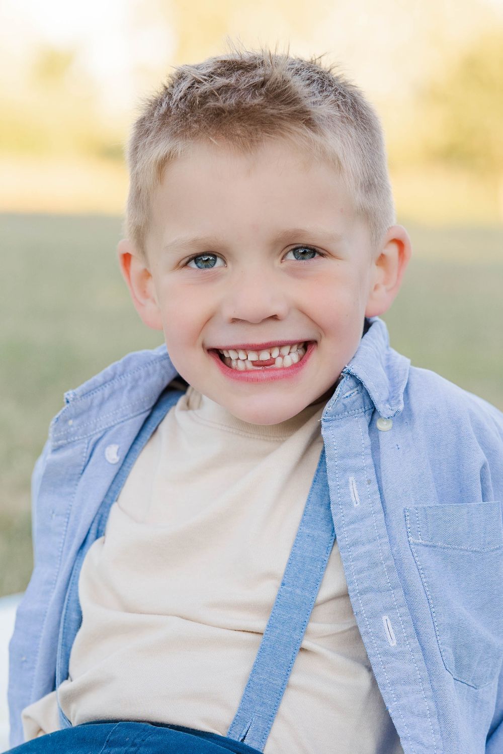 Smiling young blonde boy in a blue button-up shirt poses outdoors in natural light.