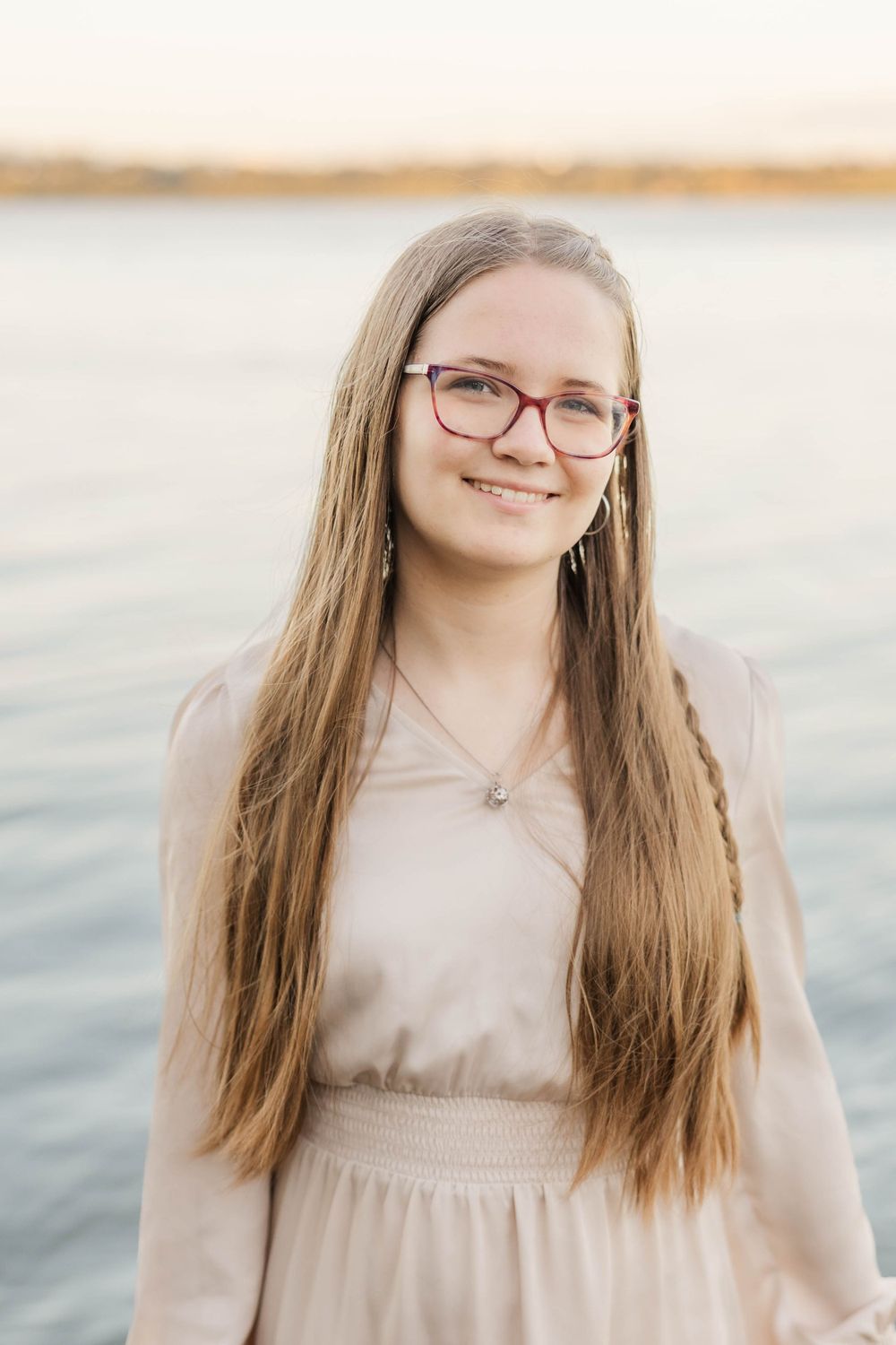 Teenage girl with long hair and red glasses smiles by a lake at sunset in a blush dress.