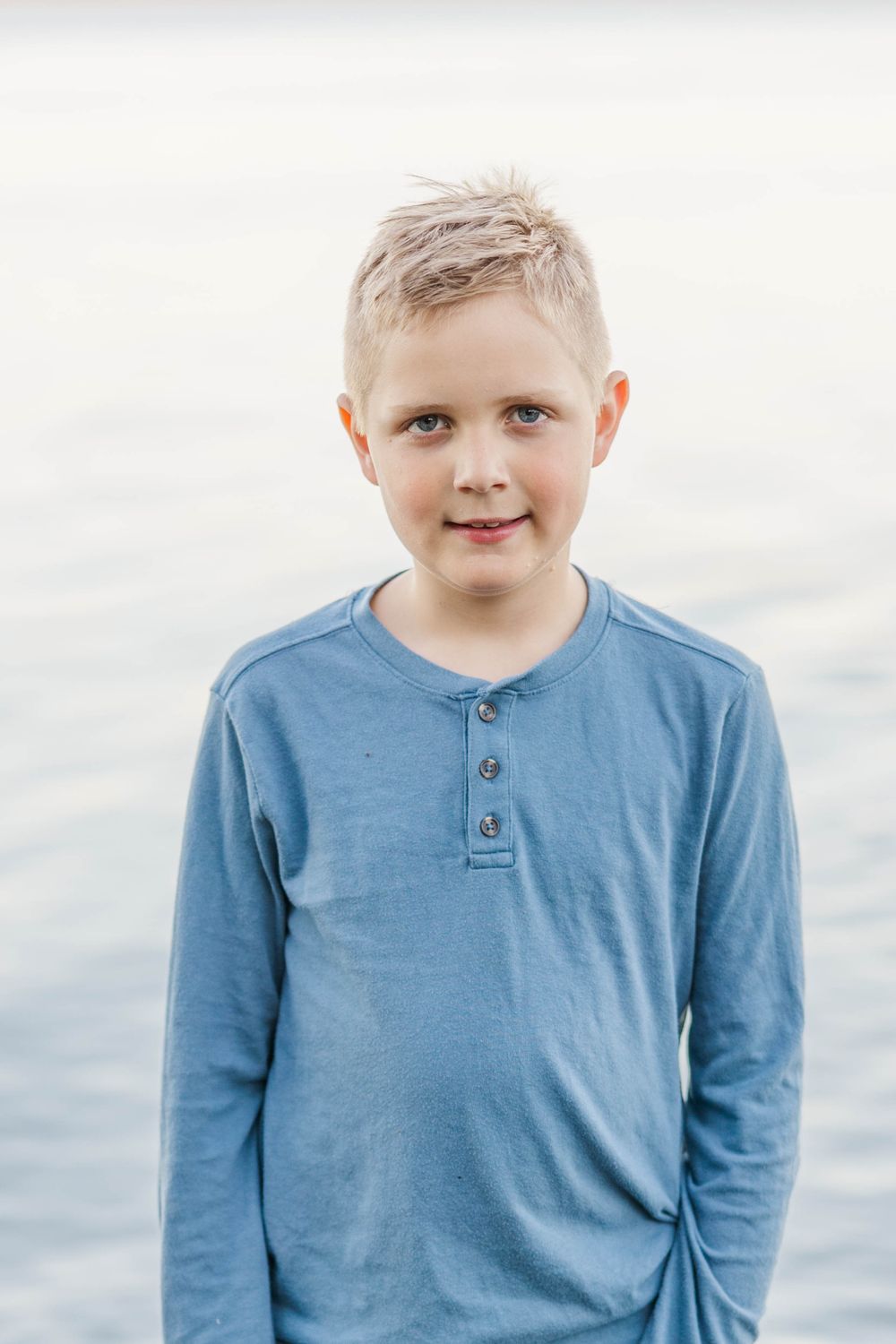 Young blonde boy in a blue henley shirt stands near water with a calm smile.