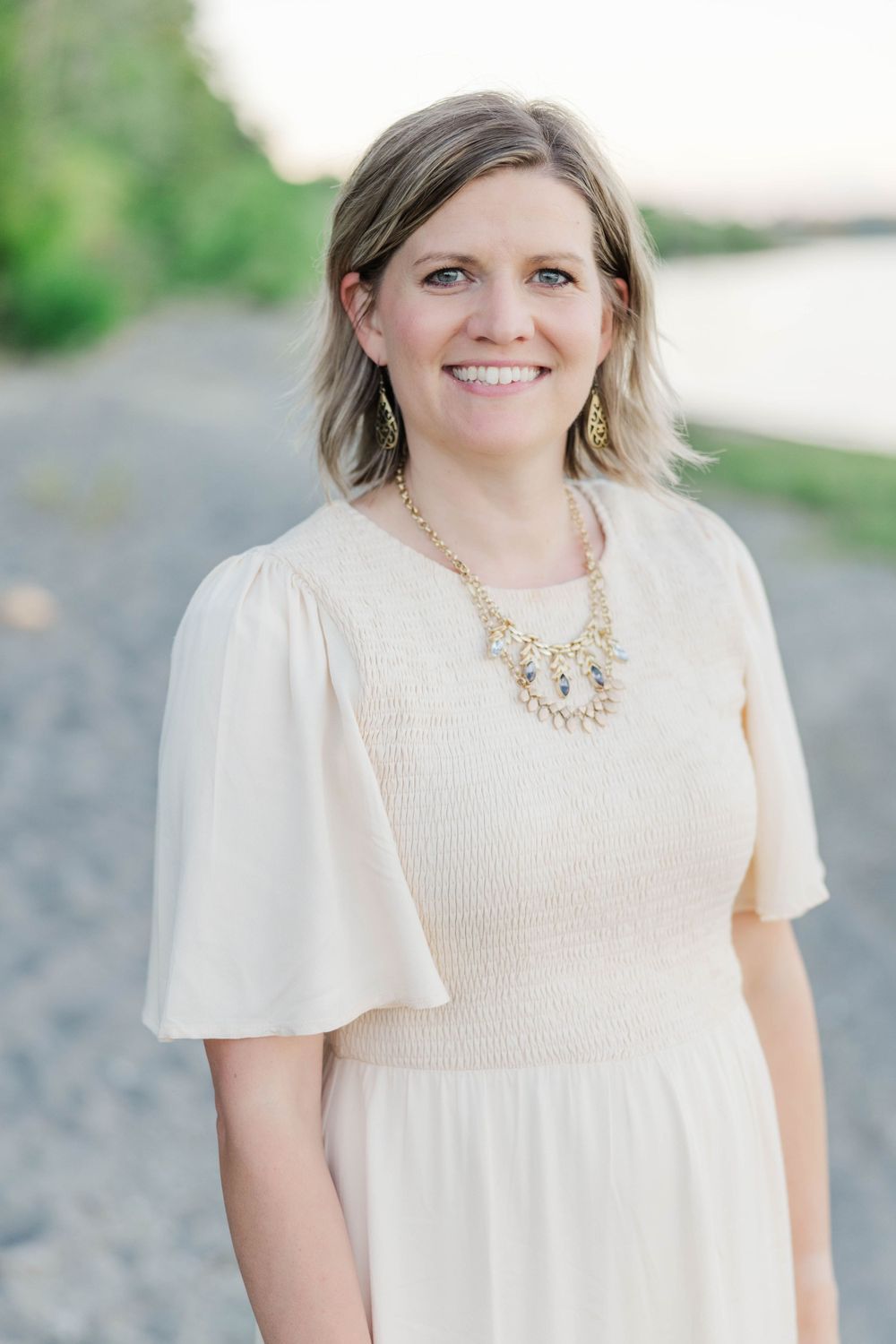 Smiling woman in cream dress with gold jewelry poses on a gravel path near a river.