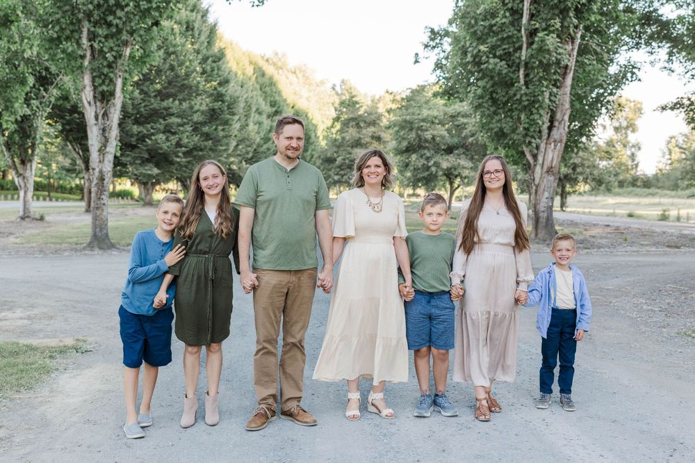 Family of seven poses together on a sunny dirt path surrounded by green trees.