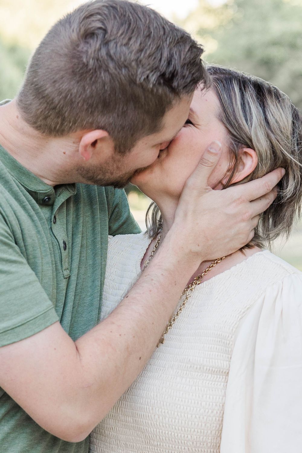Couple shares a romantic kiss outdoors surrounded by soft natural green bokeh.