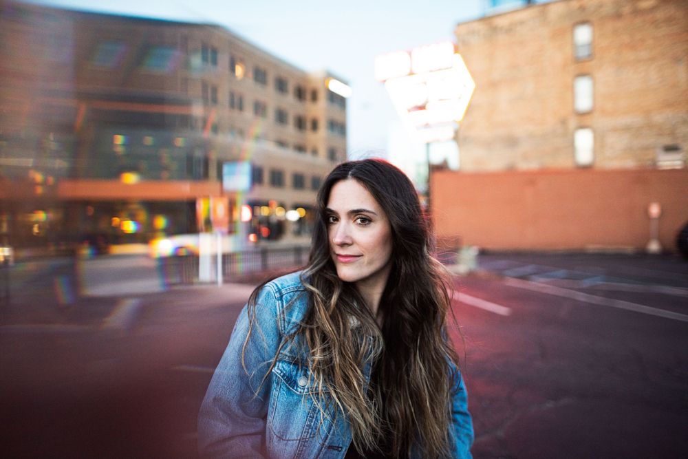 Person wearing denim jacket stands on urban street at dusk with red brick buildings in background.