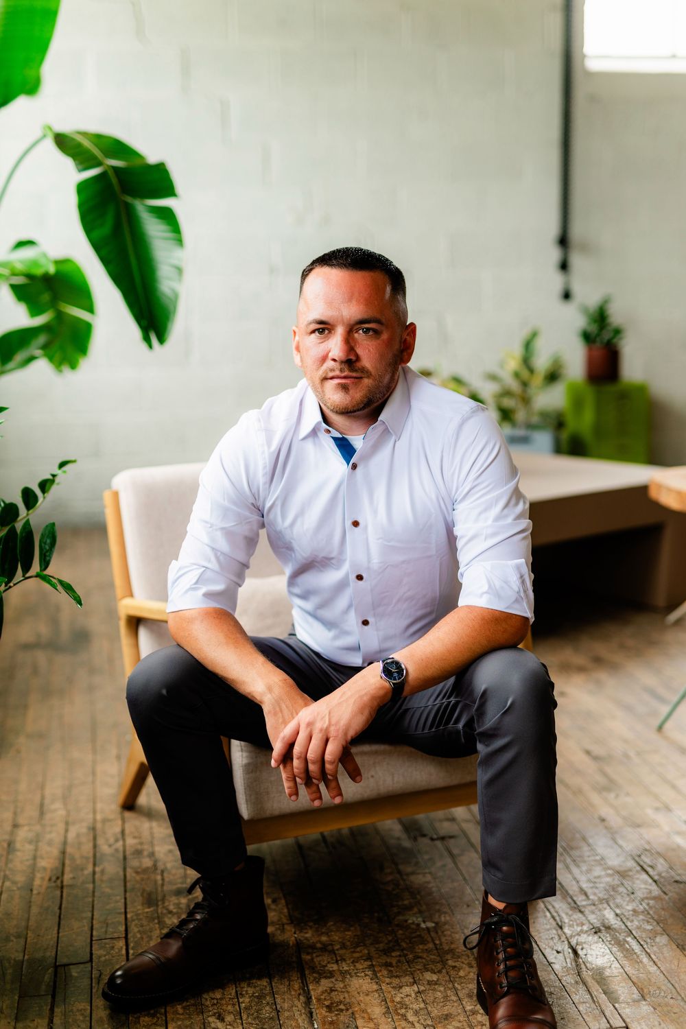 Man in white shirt and gray pants sitting confidently in a modern chair surrounded by green plants on wood floor.