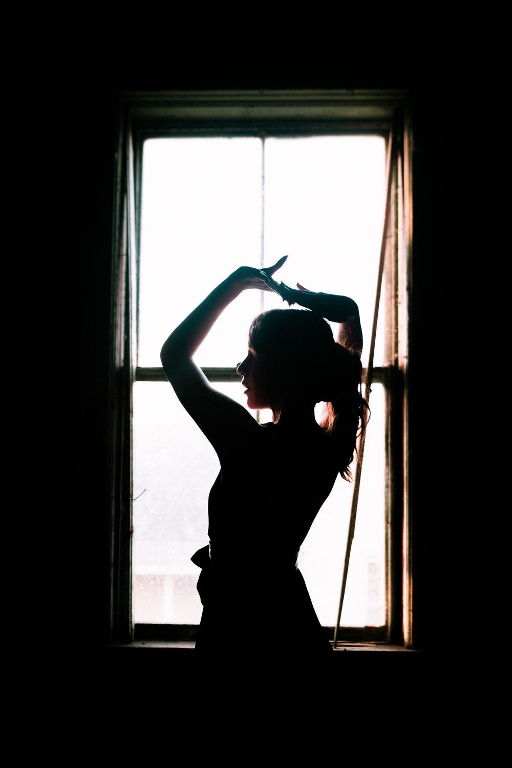 A silhouetted figure poses gracefully against a bright window, creating a dramatic backlit contrast in dark room.
