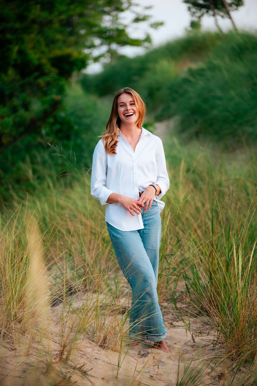 An outdoor portrait photoshoot in tall beach grass in west michigan shows a relaxed figure in a white button-down shirt and jeans.