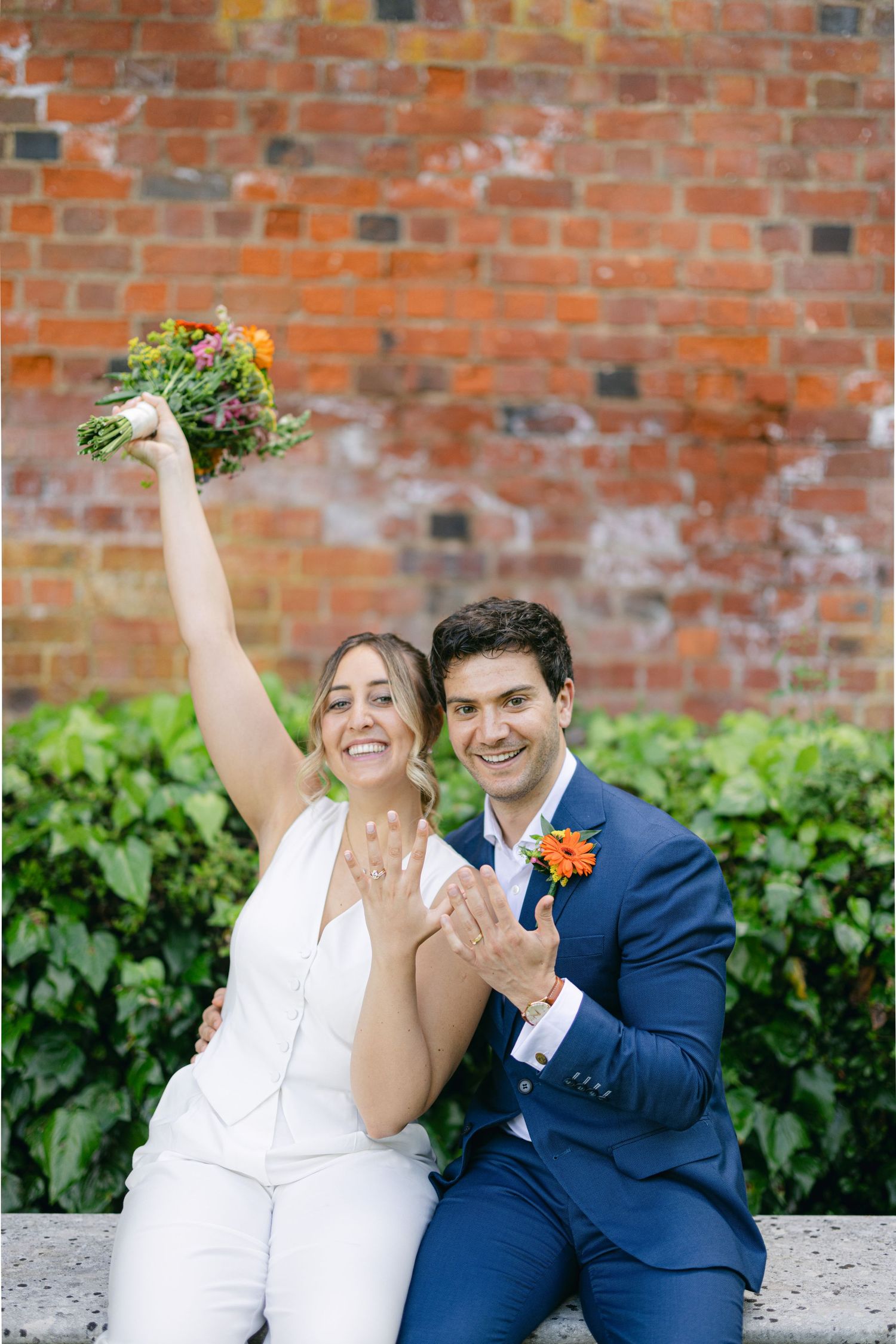 A celebratory moment between newlyweds against a brick wall, with raised bouquet and joyful expressions.
