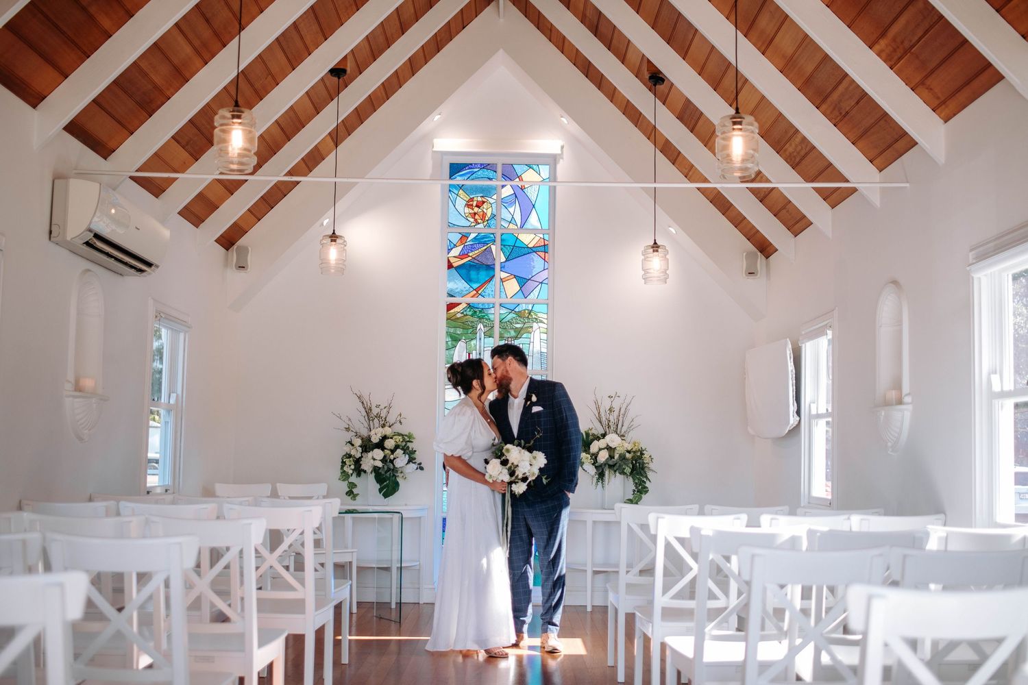 Interior of a bright white chapel with exposed wooden beams and cross-back chairs.