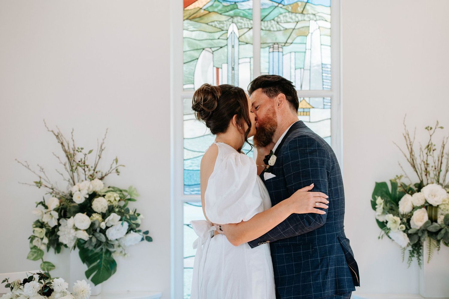 Intimate moment between newlyweds in white chapel with floral arrangements.