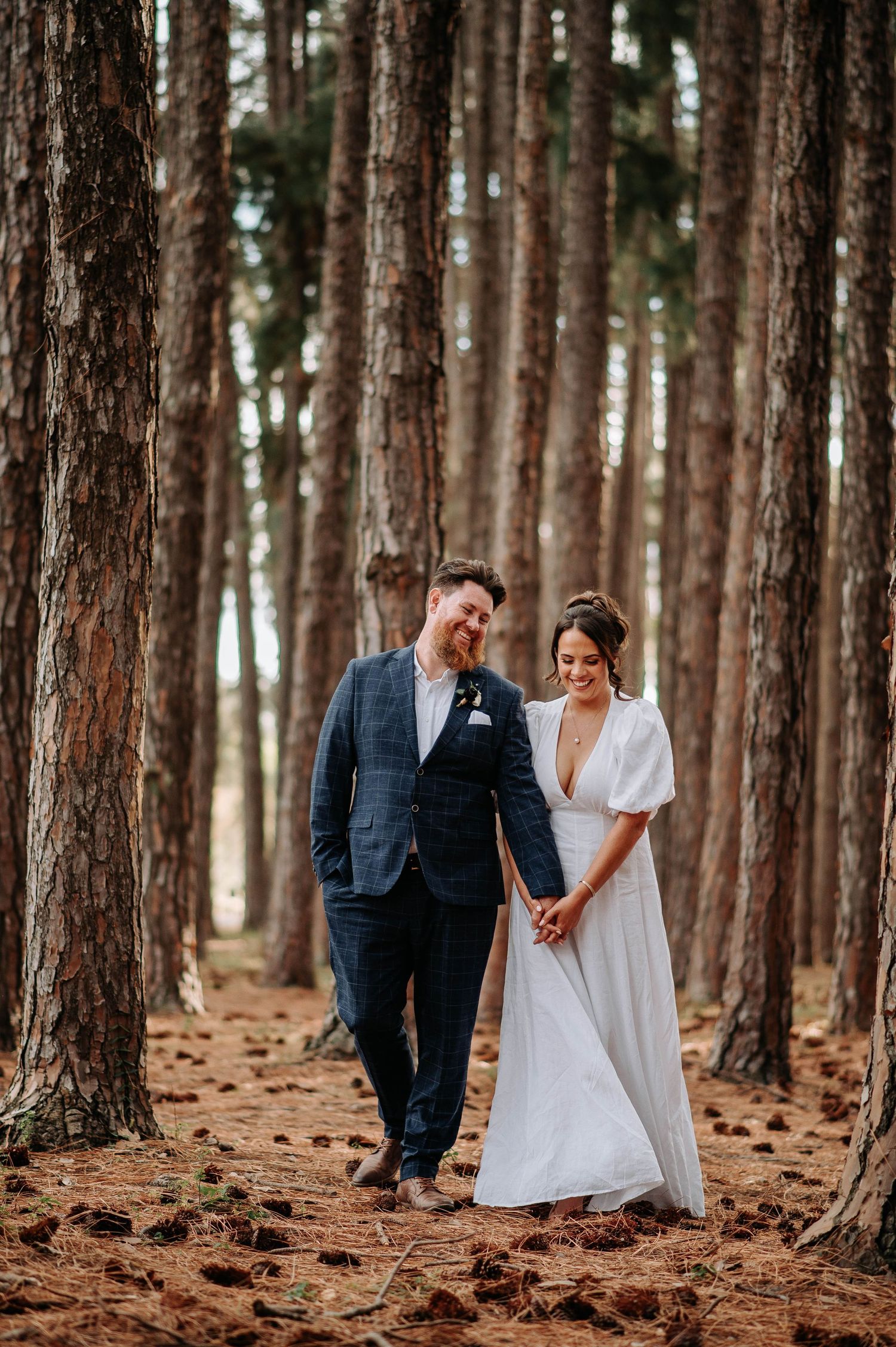 A wedding couple walking together in a serene pine forest with sunlit trees.