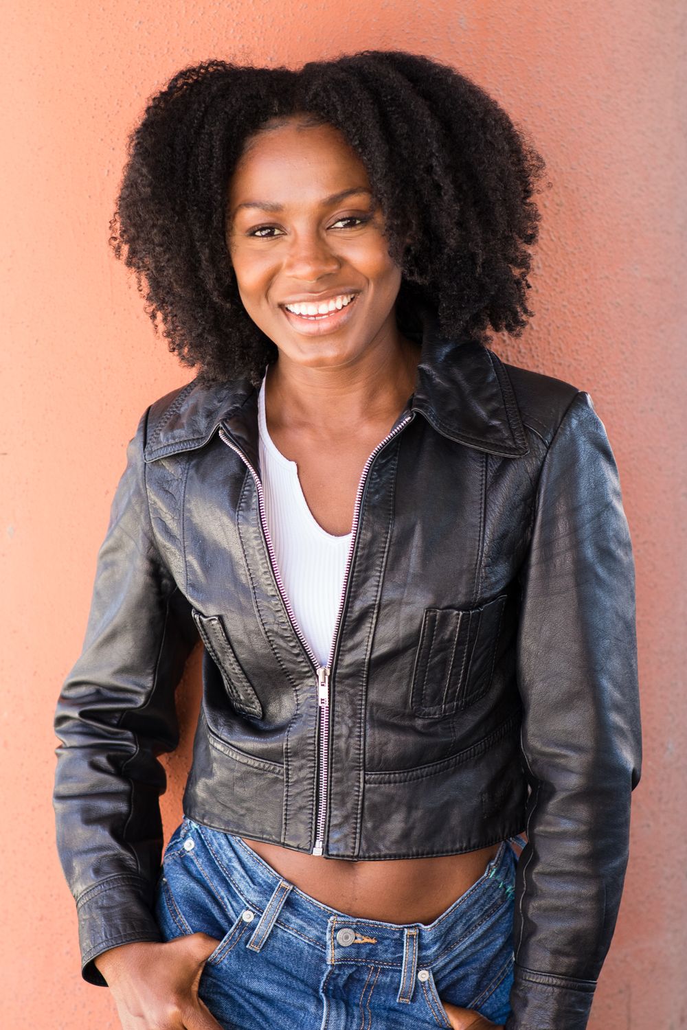 Confident woman with natural curly hair wearing leather jacket and jeans against coral background.