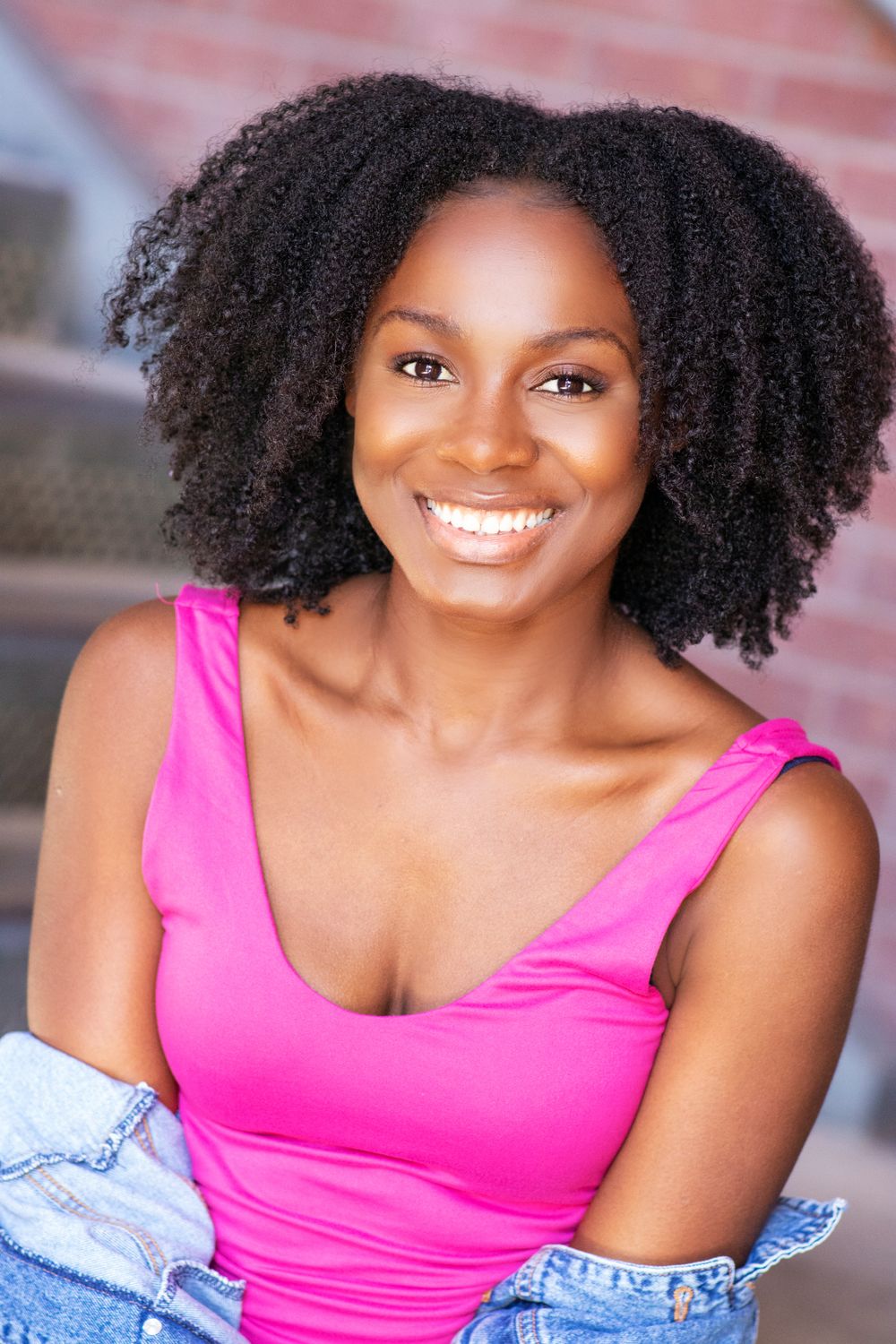 Outdoor portrait with natural curly hair wearing bright pink top and denim jacket.