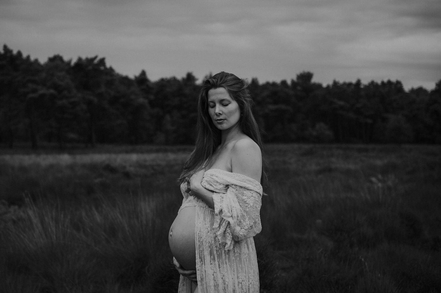 Black and white maternity portrait in a field with a lace dress against a moody sky.
