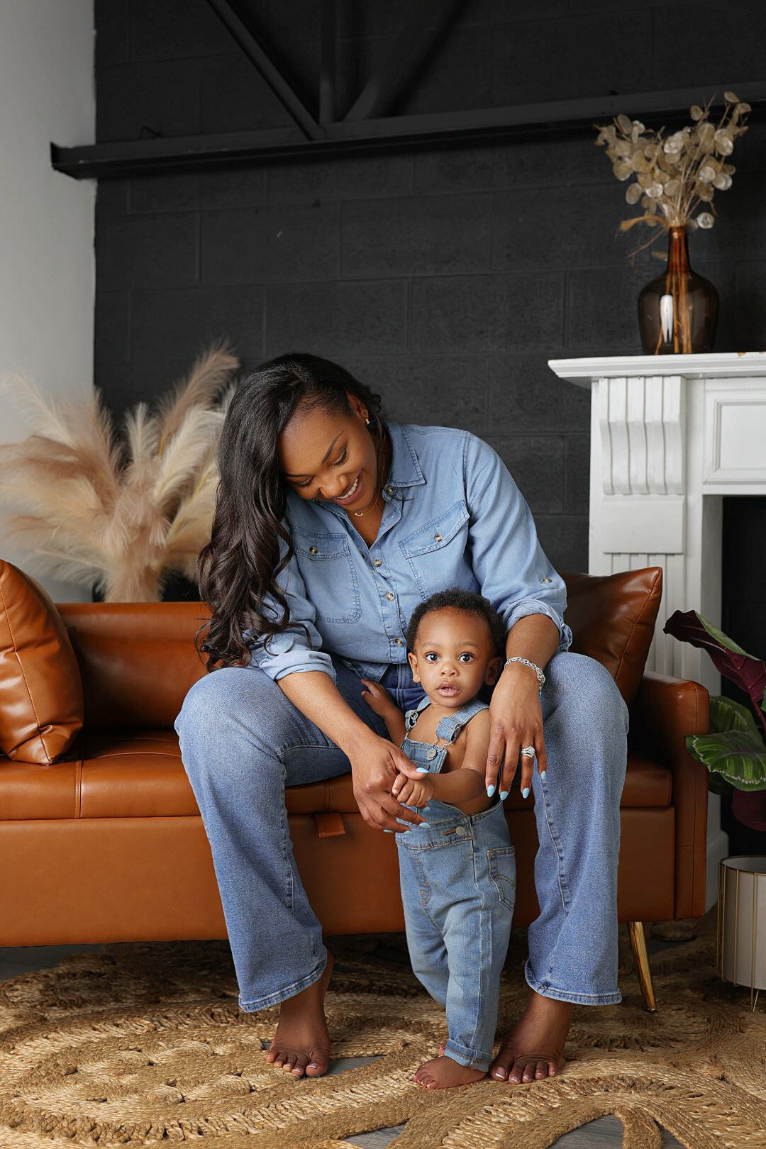 Mother in a denim outfit smiles at her toddler in matching denim while sitting barefoot on a tan leather couch in a stylish living room.